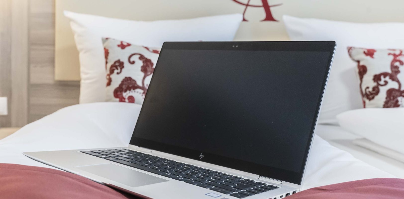 Ein Laptop liegt auf einem Bett, das mit einem roten Kissen dekoriert ist.